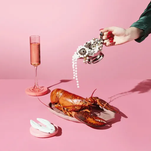 Advertising image of jewlery being poured over plate with a lobster on it with dramatic studio lighting. On Pink background.