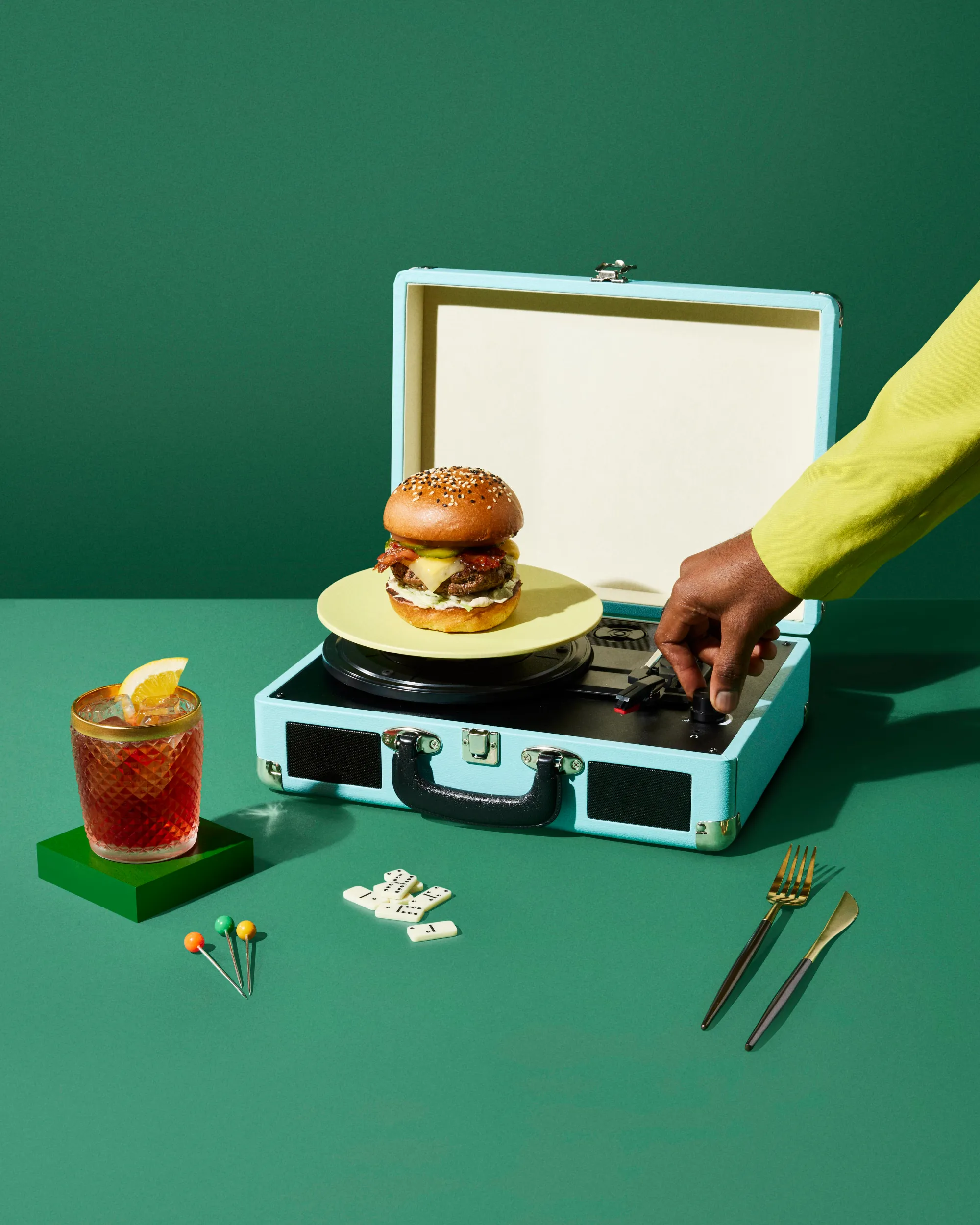 Advertising image of mans hand turning on vinyl player with a plate and burger on the player. With dramatic studio lighting. On Green background.