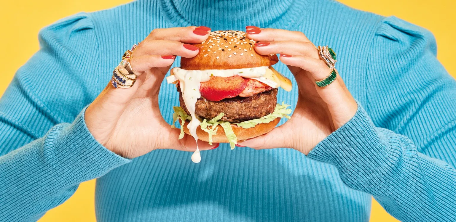 Advertising image of a luxury burger being held by female hands with bright coloured rings on. With dramatic studio lighting.
