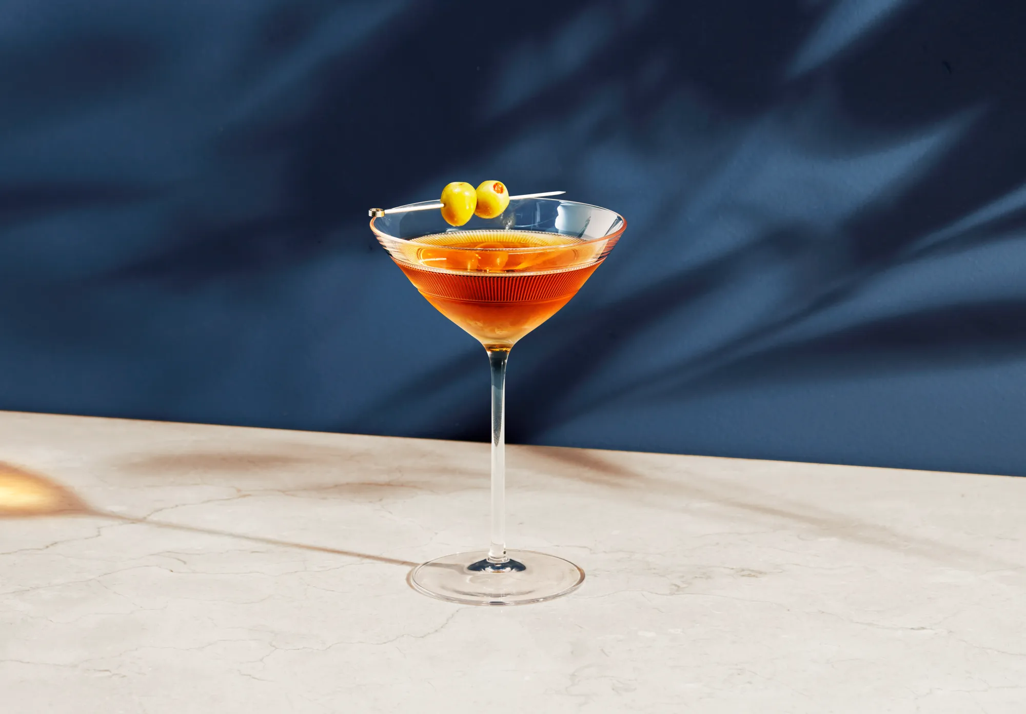still life photograph of metaxa cocktail in martini glass in evening sun. Blue background and reflection of liquid on the surface. faint shadows of branches on the background. cocktail garnished with silver pick and olives