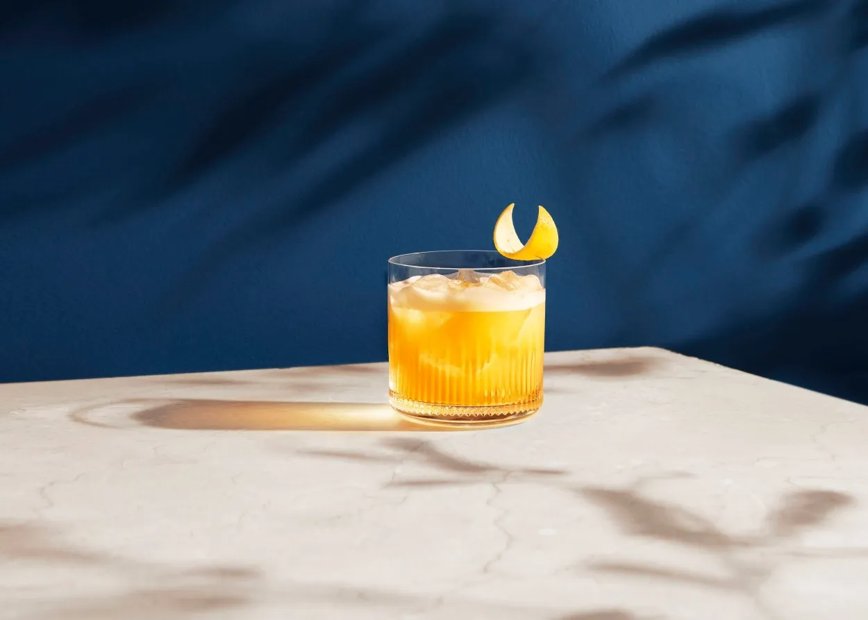 still life photograph of metaxa cocktail in a tumbler glass in evening sun. midnight blue background background and reflection of liquid on the surface. faint shadows of branches on the background