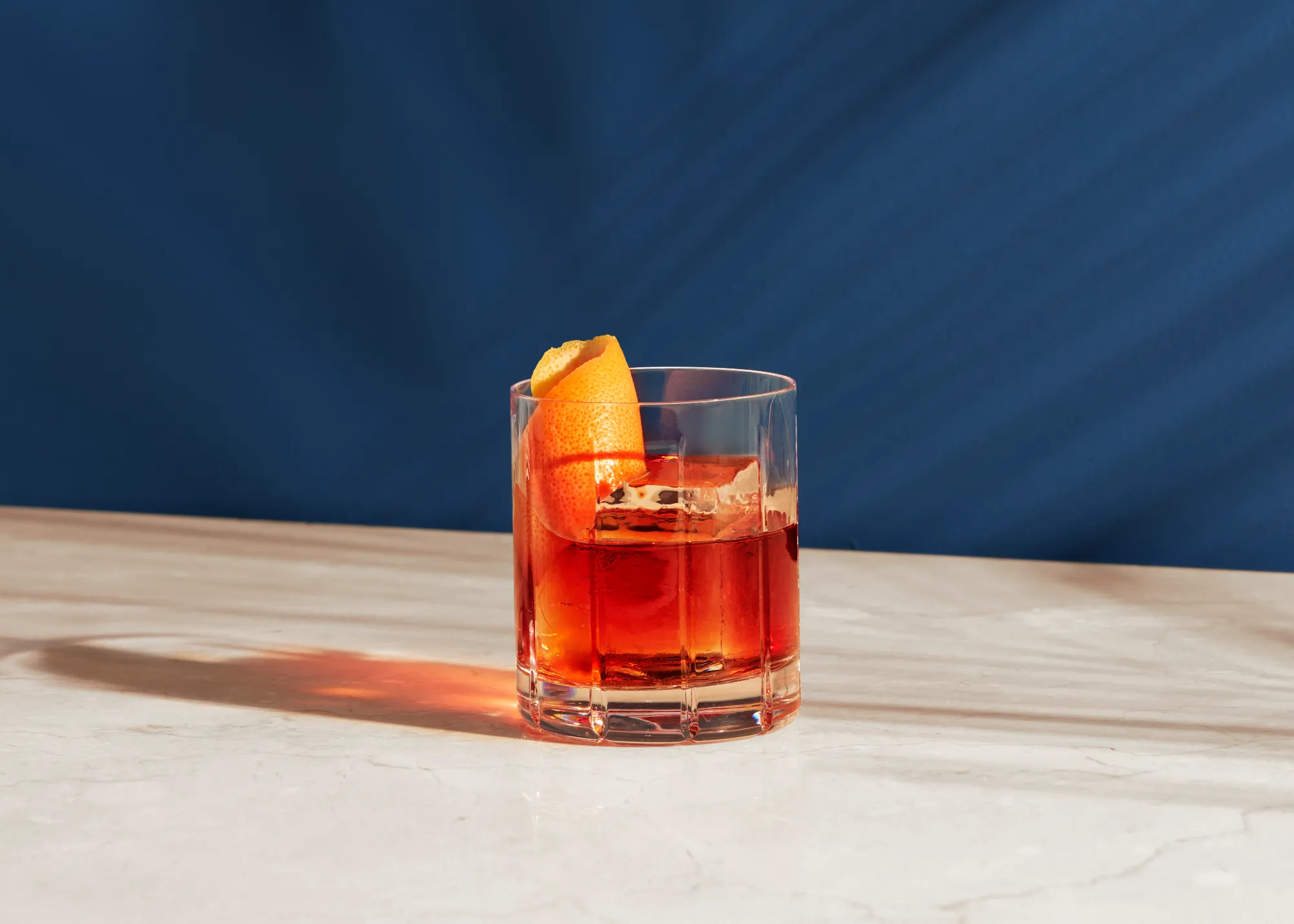 still life photograph of red metaxa cocktail in a tumbler glass in evening sun. midnight blue background background and reflection of liquid on the surface. faint shadows of branches on the background. Garnished with orange peel
