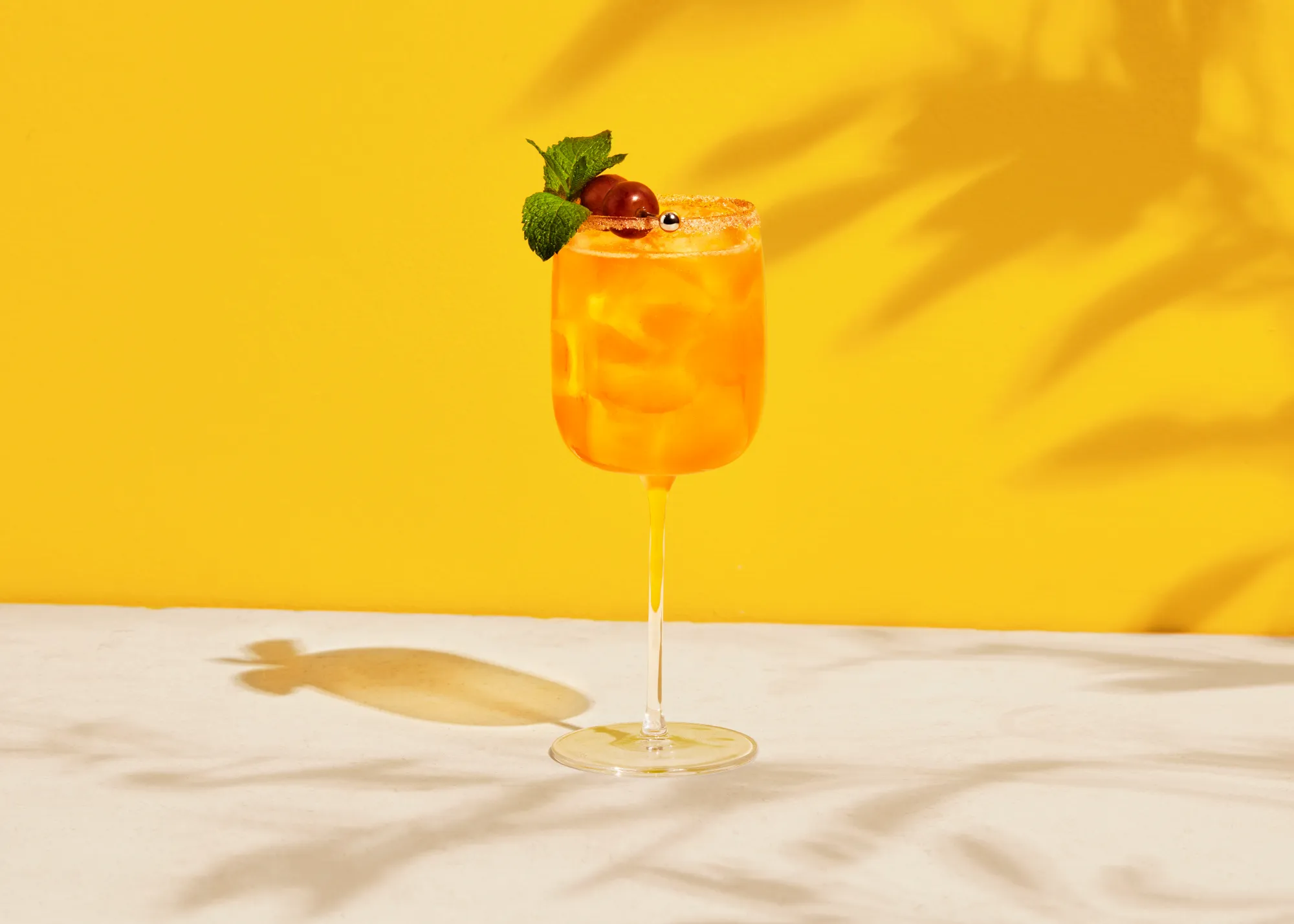 still life photograph of metaxa orange cocktail in afternoon sun sun. yellow background and reflection of liquid on the surface. faint shadows of branches on the background and surface. cocktail garnished with mint leave and cherries 