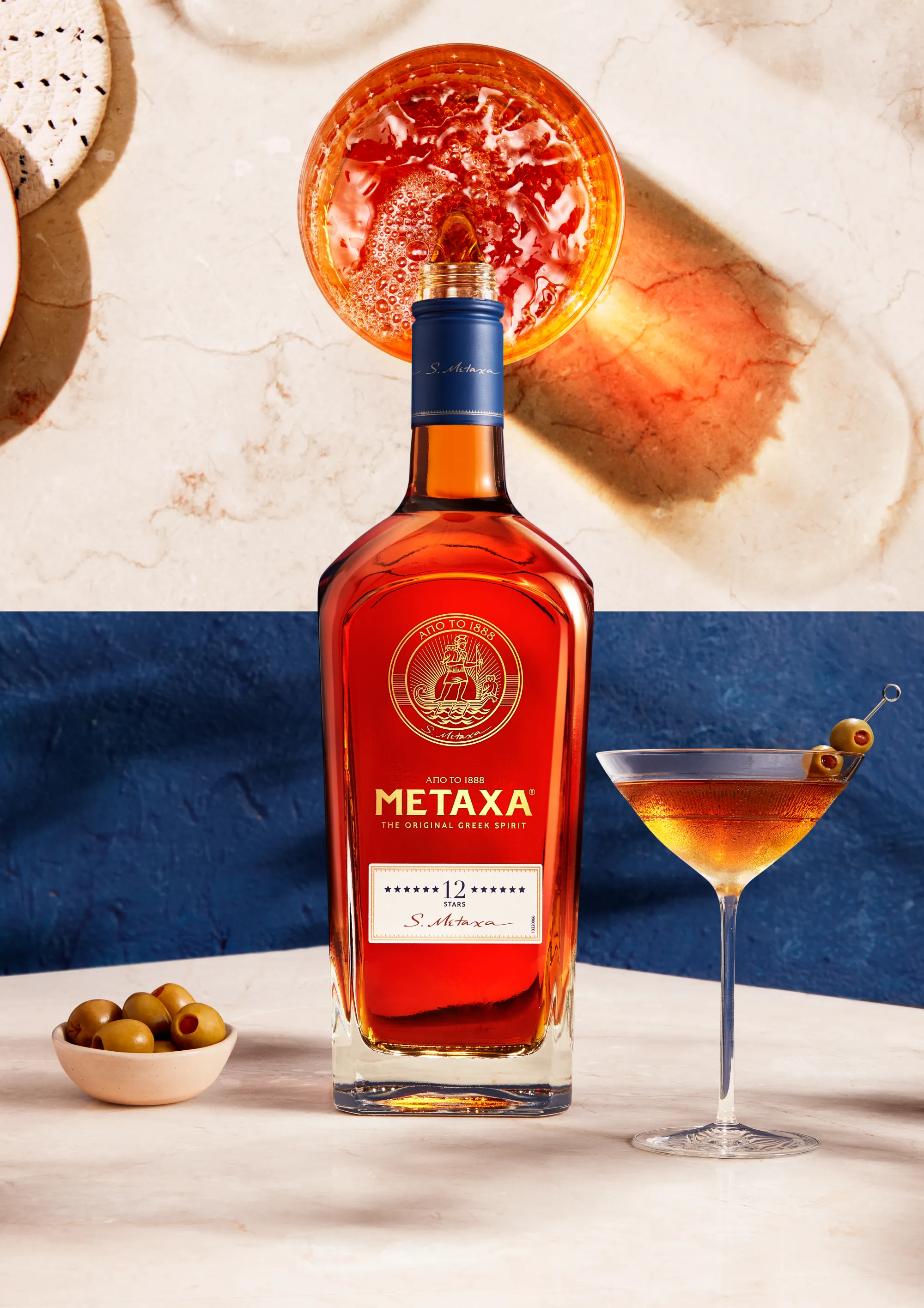 Still life photograph of Metaxa twelve star bottle on marble base with a cocktail in a martini glass next to it. The image splits half way up to show the top of the bottle pouring metaxa into glass with shadows and reflection of liquid on the surface