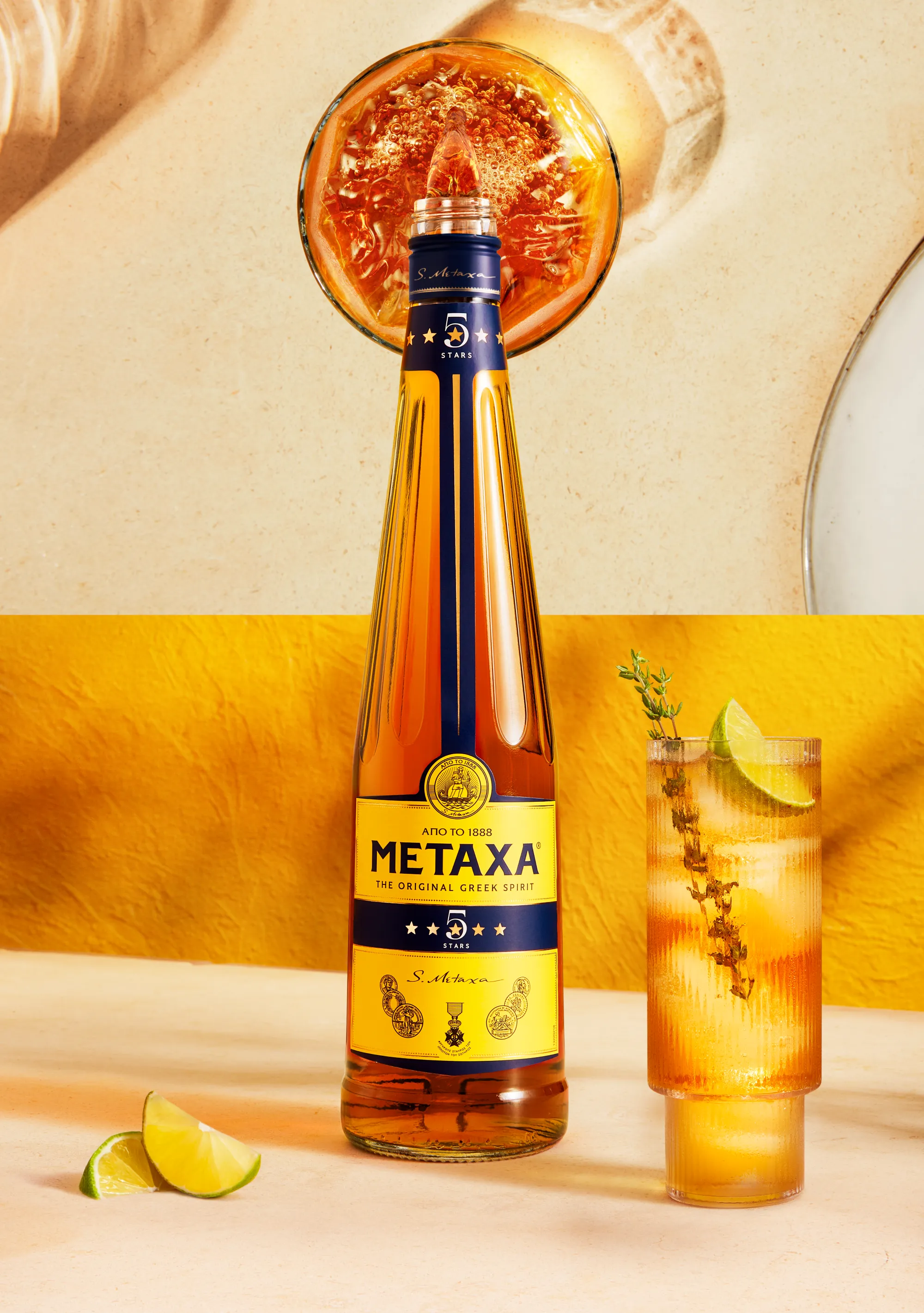 Still life photograph of Metaxa five star bottle on marble base with a cocktail in a tall glass next to it. The image splits half way up to show the top of the bottle pouring metaxa into glass with shadows and reflection of liquid on the surface