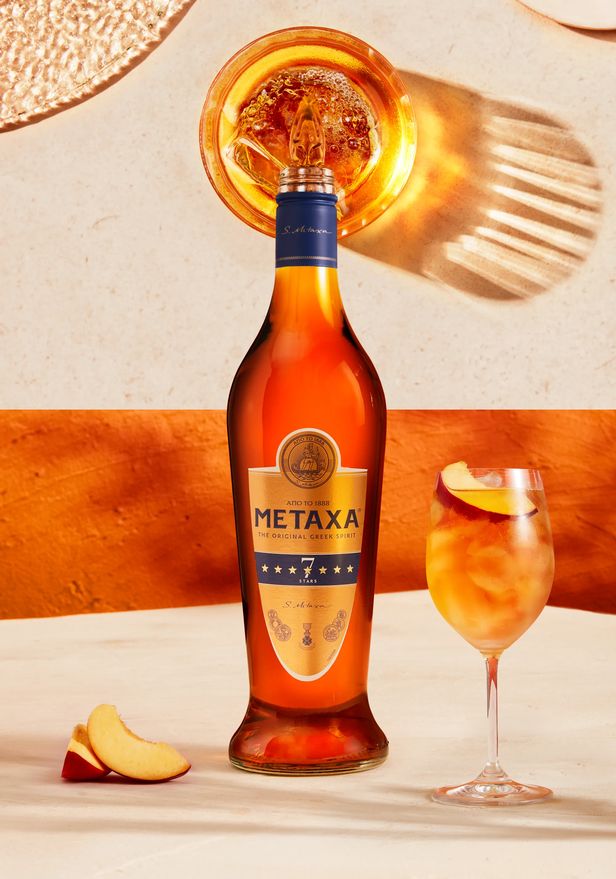 Still life photograph of Metaxa seven star bottle on marble base with a cocktail in a wine glass next to it. The image splits half way up to show the top of the bottle pouring metaxa into glass with shadows and reflection of liquid on the surface